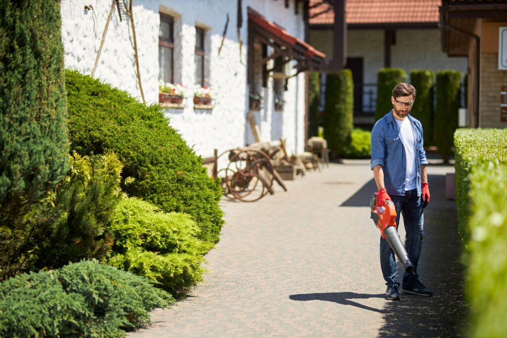 Ein Mann mit Bart, Jeanshemd und Schutzbrille nutzt einen leuchtend roten Laubbläser auf einem gepflasterten Hof. Er trägt rote Arbeitshandschuhe und konzentriert sich auf die Reinigung des Weges. Die Szene ist von üppigen grünen Hecken und Sträuchern gesäumt; im Hintergrund ist ein weißes Gebäude mit rustikalem Charme und Blumenkästen unter sonnigem Himmel zu sehen.