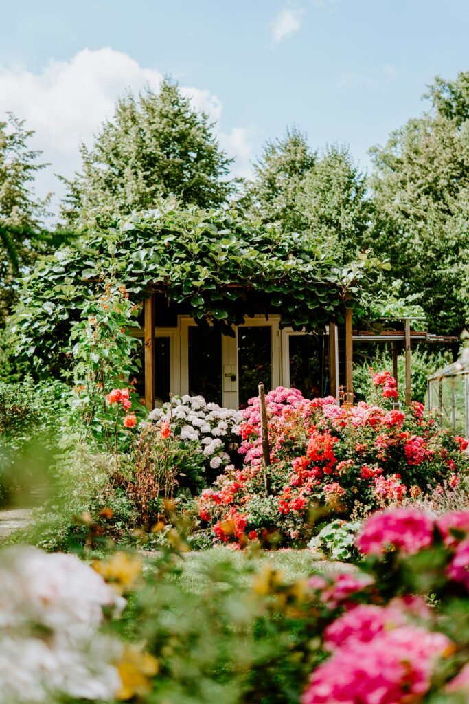 Ein idyllisches Gartenhaus, das fast vollständig von einer dicht bewachsenen grünen Pergola überrankt wird. Vor dem Häuschen erstreckt sich ein üppiges Blumenmeer aus roten, rosa und weißen Rosen sowie Hortensien. Im Hintergrund ragen hohe grüne Bäume in einen leicht bewölkten, hellblauen Himmel, während im Vordergrund weitere Blüten unscharf angedeutet sind.
