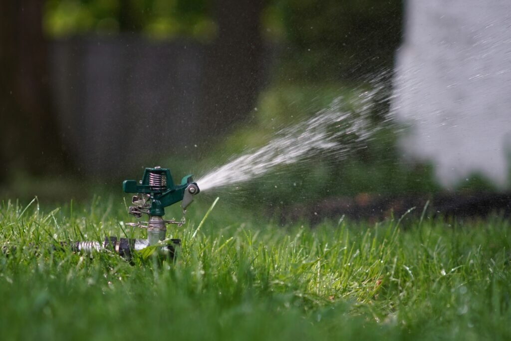 Nahaufnahme eines grünen Impulsregners, der in einem saftig grünen Rasen steht und einen fächerförmigen Wasserstrahl versprüht. Die Wassertropfen glitzern im Licht vor einem unscharfen, dunkleren Hintergrund aus Gartenbepflanzung.