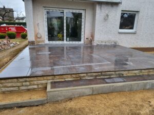 Eine neu verlegte, erhöhte Terrasse mit großen, dunkelbraun glänzenden Fliesen. Die Terrassenkante besteht aus hellen Mauersteinen. Das Haus im Hintergrund befindet sich noch in der Sanierung, erkennbar an der abgebrochenen Fassade um die neue Glasschiebetür und einem roten Schuttcontainer im Garten.