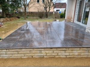 Eine erhöhte Terrasse mit nassen, dunkelbraunen Fliesen, die vor einem Wohnhaus verlegt wurden. Die Terrassenkante besteht aus hellen Mauersteinen. Ein Handwerker in Arbeitskleidung ist am linken Rand dabei, letzte Arbeiten an den Fliesen vorzunehmen. Im Hintergrund ist ein Garten mit einem Holzzaun und einem weiteren Haus unter bewölktem Himmel erkennbar.