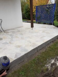 Eine erhöhte Terrasse aus hellen, polygonalen Natursteinplatten in Weiß- und Beigetönen. Ein dunkler Holzpfosten stützt das Vordach, während im Hintergrund ein blaues Rankgitter steht. Im Vordergrund ist ein Handwerker beim Verputzen des Terrassensockels zu sehen.