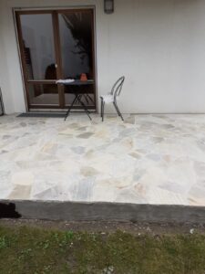 Eine fertiggestellte Terrasse aus hellen Polygonalplatten aus Quarzit oder Naturstein in verschiedenen Weiß- und Beigetönen. Auf der Fläche stehen ein kleiner runder Metalltisch und ein Stuhl vor einer großen Glasschiebetür.