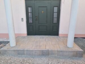 Neu gestalteter Hauseingang mit einem Podest aus großformatigen, sandfarbenen Steinplatten und massiven Blockstufen. Zwei weiße Säulen rahmen die dunkelgrüne Haustür ein.