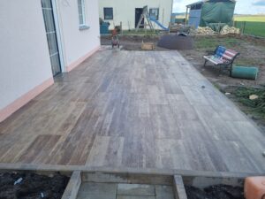 Eine neu angelegte, großflächige Außenterrasse mit graubraunen Fliesen in Holzoptik, angrenzend an eine helle Hauswand mit einer Balkontür. Im unfertigen Gartenbereich dahinter steht eine Parkbank mit USA-Flaggenmuster.