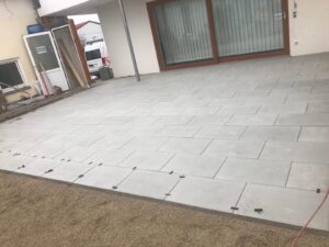 Blick auf eine großflächige, erhöhte Terrasse aus rechteckigen, grauen Steinplatten, die vor einem Wohnhaus verlegt werden. An den offenen Fugen der vorderen Platten sind schwarze Abstandshalter zu sehen, die den laufenden Verlegeprozess verdeutlichen. Die Terrasse grenzt an einen unfertigen Gartenbereich mit Sanduntergrund. Im Hintergrund sind große Glasschiebetüren mit Holzrahmen, eine weiße Hauswand und ein weißer Lieferwagen erkennbar.