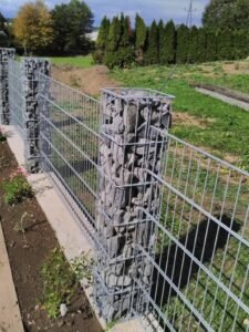 Ein modern gestalteter Gartenzaun bestehend aus grauen Doppelstabmatten und schmalen Gabionen-Säulen, die mit dunklen Natursteinen gefüllt sind. Der Zaun ist auf einem Betonsockel montiert, der an ein bepflanztes Beet grenzt. Im Hintergrund sind eine grüne Wiese und eine hohe Thuja-Hecke unter leicht bewölktem Himmel zu sehen.