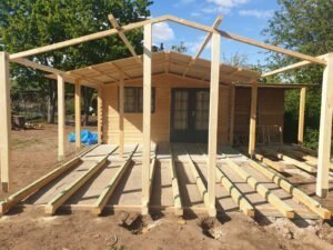 Ein Gartenhaus aus hellem Holz im Bauzustand. Vor dem Haus ist das hölzerne Grundgerüst für eine große Terrassenüberdachung und ein Balkenfundament für den Bodenbelag zu sehen. Das Haus hat eine dunkle Doppeltür und Sprossenfenster.
