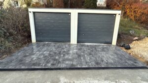 Eine gepflasterte Garagenauffahrt vor zwei dunkelgrauen Sektionaltoren mit weißem Rahmen. Das Pflaster besteht aus rechteckigen Steinen in melierten Grautönen. Im Vordergrund verläuft eine Entwässerungsrinne entlang der gesamten Breite. Die Garagen sind in einen Hang gebaut, der von herbstlichen Sträuchern und Bäumen umgeben ist.