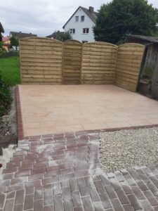 Kombiniertes Pflaster- und Holzbauprojekt: Eine neue Terrasse aus hellen Steinplatten ist mit sauberen Rändern eingefasst. Dahinter wurde eine stabile Sichtschutzwand aus hellen, gewölbten Holzelementen errichtet. Der Zugangsbereich ist mit roten Pflastersteinen und Schotter vorbereitet.