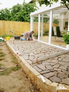 Ein Handwerker klopft mit einem Gummihammer Naturstein-Polygonalplatten in ein Mörtelbett ein. Die Terrasse wird direkt vor einem modernen, weißen Wintergarten errichtet.