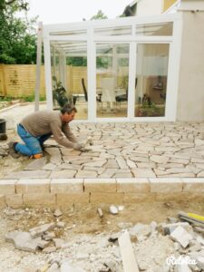 Ein Handwerker kniet auf einer Baustelle und verlegt in präziser Handarbeit Polygonalplatten aus Naturstein auf einer Terrasse vor einem modernen Wintergarten.