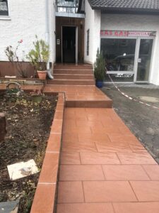Ein frisch sanierter und gefliester Eingangsbereich eines Wohn- und Geschäftshauses. Ein langer, barrierefreier Weg aus terracottafarbenen Fliesen führt über ein Podest und drei Stufen direkt zur Haustür. Rechts davon ist ein Ladeneingang (Honda, Gas Gas) mit großen Glasfronten zu sehen. Ein kleines Beet grenzt links an den Weg.