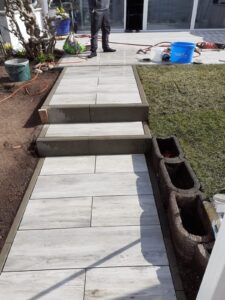 Eine im Bau befindliche Gartentreppe aus hellgrauen Steinplatten in Holzoptik. Der Weg führt über drei Stufen nach oben. Rechts davon befindet sich eine Reihe unbepflanzter Pflanzkästen, links aufgeschüttete Beeterde. Zwei helle Blockstufen bilden den Abschluss. Im Hintergrund steht eine Person und es ist Baustellen-Equipment zu sehen.