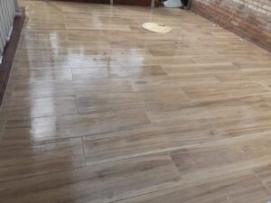 Eine großflächig geflieste Terrasse mit hellbraunen Bodenplatten in täuschend echter Holzoptik. Die glänzende Oberfläche spiegelt das Licht wider und grenzt rechts an eine rustikale Backsteinmauer.