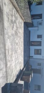 Eine gepflasterte Einfahrt vor einem weißen Wohnhaus, verlegt mit rechteckigen Pflastersteinen in verschiedenen Grautönen. Die Fläche ist sauber eingefasst und führt direkt zu den Hauseingängen. Im Hintergrund sind ein Holzzaun, Mülltonnen und die Fassade des Gebäudes bei hellem Tageslicht zu sehen.