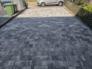 Ansicht einer frisch gepflasterten, breiten Hauseinfahrt. Der vordere Bereich ist mit modernen anthrazit-melierten Pflastersteinen belegt, während der hintere Teil, der zur Straße führt, mit hellgrauen Pflastersteinen abgesetzt ist. Rechts befindet sich ein Jägerzaun, links Mülltonnen.