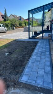 Ein neu gepflasterter Gartenweg aus blaugrauen, rechteckigen Steinen, der entlang eines Hauses zu einer modernen Außentreppe mit Glasüberdachung führt. Der angrenzende Gartenbereich ist noch unbefestigt. Im Hintergrund sind eine gepflasterte Einfahrt, eine Straße mit parkenden Autos und weitere Wohnhäuser unter strahlend blauem Himmel zu sehen.