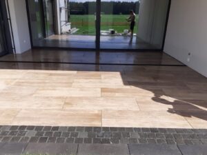 Moderne Terrassengestaltung mit großformatigen Platten in heller Holzoptik, kombiniert mit einer dunklen Naturstein-Pflasterung als Abschlusskante. Im Hintergrund spiegelt sich der Garten in einer großen Glasfront.