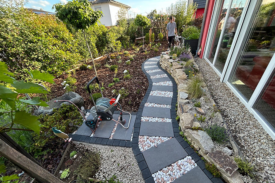 Ein geschwungener Gartenweg aus anthrazitfarbenen Betonplatten und hellen Kieselsteinen führt durch einen gepflegten Garten mit Rindenmulch, Staudenbeeten und einer Natursteinmauer direkt am Haus.