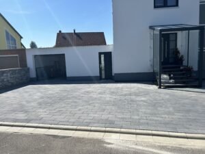 Eine weitläufige, modern gepflasterte Hauseinfahrt mit grauen, rechteckigen Steinen. Das Pflaster grenzt direkt an ein weißes Wohnhaus mit verglastem Eingangsbereich und eine angrenzende Garage. Die gesamte Fläche ist sauber mit Bordsteinen zur Straße hin abgeschlossen und wird von hellem Sonnenlicht beleuchtet.