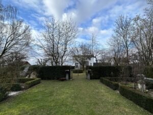 Blick durch einen weitläufigen Garten mit einer zentralen Rasenfläche, die von niedrigen Buchsbaumhecken und Beeten flankiert wird. Eine hohe, dunkle Hecke mit einem mittigen Durchgang unterteilt das Grundstück. Dahinter sind laublose Bäume und ein weißes Wohnhaus unter einem bewölkten blauen Himmel erkennbar.