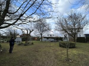 Blick über einen weitläufigen Garten im frühen Frühling. Links steht ein Mann in Arbeitskleidung und schaut in den Garten. Zu sehen sind viele laublose Bäume, ein Hühnerstall, Hochbeete und im Hintergrund Wohnhäuser unter bewölktem Himmel.
