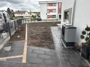 Ein gepflasterter Weg aus großformatigen, dunkelgrauen Steinplatten führt entlang einer weißen Hauswand mit einer installierten Wärmepumpe. Der Weg wird durch eine Kante aus helleren Steinen von einer vorbereiteten, erdigen Gartenfläche abgegrenzt. Im Hintergrund sind ein grauer Metallzaun, ein weißer Kleintransporter und weitere Wohnhäuser unter bewölktem Himmel zu sehen.