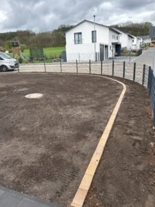 Eine großflächige, vorbereitete Erdfläche im Garten eines Neubaus, die mit einer geschwungenen Kante aus hellen Pflastersteinen eingefasst ist. Ein dunkler Doppelstabmattenzaun begrenzt das Grundstück zur Straße hin. Im Hintergrund sind moderne weiße Wohnhäuser, eine hügelige Landschaft mit Bäumen und ein bewölkter Himmel zu sehen.