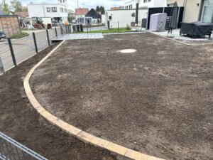 Eine großflächige, vorbereitete Gartenfläche mit dunkler Erde, die für eine neue Rasenanlage oder Bepflanzung bereit ist. Ein geschwungener Randabschluss aus hellen Pflastersteinen trennt die Fläche vom Zaun ab. Im Hintergrund sind moderne Wohnhäuser, eine gepflasterte Terrasse mit Gartenmöbeln und ein grauer Metallzaun zu sehen.