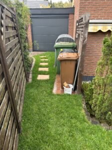 Gepflegter Gartenweg mit Schrittsteinen, der zu einer modernen Garage führt. Rechts sind ordentlich aufgereihte Mülltonnen (braun, schwarz, grün) unter einem Metallunterstand zu sehen. Die Mülltonnen stehen auf Pflastersteinen und sind von einer Hecke und einem Holzzaun umgeben. Im Hintergrund ein rotes Backsteinhaus mit einer gelben Markise.