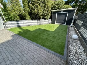 Ein gepflegter Hinterhof-Garten, aufgenommen bei Sonnenschein. Links ist eine graue Pflasterterrasse, die zu einer sauberen, rechteckigen Kunstrasenfläche führt. Ein grauer Lamellenschutzzaun mit weißen Akzenten umgibt den Garten. Im Hintergrund steht ein modernes, dunkelgraues Gartenhaus mit Rolltor und weißem Dachrand, umgeben von dichten grünen Thuja-Bäumen. Rechts verläuft ein Kiesbeet mit zwei runden, weißen Kugelleuchten.