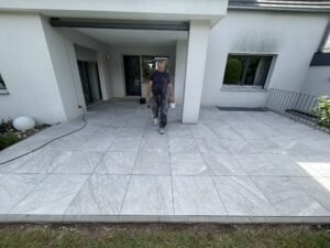 Nahaufnahme eines Fliesenlegers, der über eine frisch verlegte Terrasse mit großen grauen Fliesen geht. Die gleichmäßigen Fugen und der saubere Abschluss an der Hauswand zeigen die fachgerechte Arbeit. Baueimer und Werkzeug sind im Hintergrund zu sehen.