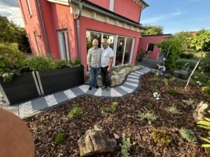 Zwei lächelnde Männer, ein Kunde und ein Handwerker, stehen Arm in Arm auf einem neuen, geschwungenen Gartenweg vor einem lachsfarbenen Wohnhaus. Der Weg ist mit rechteckigen grauen Pflastersteinen und hellem Zierkies verlegt, eingefasst von Natursteinen und Rindenmulch-Beeten. Rechts ist ein modernes dunkles Hochbeet sichtbar. Die Szene ist bei hellem Sonnenlicht aufgenommen, mit Vegetation und einem Baumstumpf im Vordergrund.