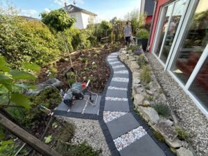 Ein geschwungener Gartenweg aus großen, grauen Pflastersteinen, deren Zwischenräume mit hellem Zierkies gefüllt sind. Der Weg führt durch einen üppigen Garten mit Beeten und einer Mauer aus rustikalen Natursteinen. Links neben dem Weg ist ein Gartenschlauch auf einem Wagen platziert. Die Szene liegt vor einem Haus mit großen Glasschiebetüren.