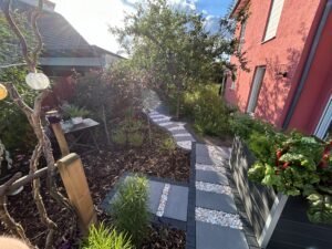 Ein geschwungener Gartenweg aus großen, grauen Steinplatten, deren Zwischenräume mit hellem Zierkies gefüllt sind. Der Weg führt durch einen üppig bepflanzten Garten mit Rindenmulch-Beeten vorbei an einem dunklen Hochbeet und einer rötlichen Hausfassade. Im Hintergrund spendet ein großer Laubbaum Schatten, während die Szene von hellem Sonnenlicht beleuchtet wird.