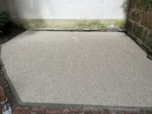 Eine exakt abgezogene und geebnete Fläche aus feinem, hellem Splitt, die als Vorbereitung für Pflasterarbeiten dient. Die Fläche ist mit dunklen Randsteinen eingefasst und grenzt an eine helle Hauswand sowie eine rustikale Natursteinmauer. Im Vordergrund ist ein schmaler Streifen mit roten Ziegelsteinen erkennbar.