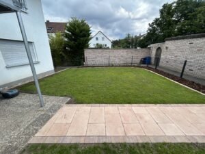 Blick auf eine gepflegte, rechteckige Rasenfläche, die im Vordergrund von einem Weg aus hellen, lachsfarbenen Terrassenplatten begrenzt wird. Der Rasen ist mit einer Kante aus Rindenmulch eingefasst und wird durch einen dunklen Metallzaun sowie eine helle Ziegelmauer vom Nachbargrundstück getrennt. Links ist die weiße Fassade eines Wohnhauses mit geschlossenem Rollladen und ein Mähroboter zu sehen.