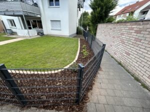 Blick auf eine gepflegte Gartenanlage mit einer Rasenfläche, die durch eine geschwungene Kante und ein Beet mit Rindenmulch eingefasst ist. Ein anthrazitfarbener Doppelstabmattenzaun trennt den Garten von einem angrenzenden, gepflasterten Gehweg und einer hellen Ziegelmauer. Im Hintergrund ist die weiße Fassade eines Mehrfamilienhauses mit Balkonen und Terrassen unter einem leicht bewölkten Himmel zu sehen.