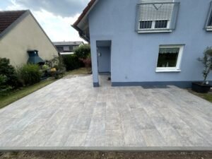 Eine fertiggestellte, großflächige Terrasse mit hellgrau melierten Steinplatten, die vor einem blau gestrichenen Wohnhaus verlegt wurden. Die Terrasse ist mit einer grauen Steinkante sauber zum Garten hin abgeschlossen. Im Hintergrund sind ein angrenzendes gelbes Gebäude mit einem Außenkamin, eine Rasenfläche sowie Sträucher unter einem leicht bewölkten Himmel zu sehen.