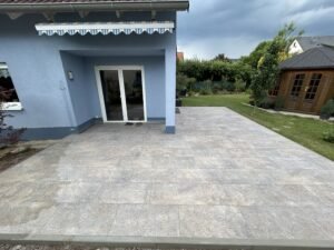 Gemini hat gesagt Alt-Text (Standard): Eine weitläufige, ebenerdige Terrasse mit großformatigen, hellgrau melierten Steinplatten vor einem blau gestrichenen Wohnhaus. Die Terrasse schließt direkt an eine gläserne Terrassentür an und wird von einer Markise beschattet. Im Hintergrund ist ein gepflegter Garten mit Rasenfläche und einem hölzernen Gartenhaus unter einem bewölkten Himmel zu sehen.