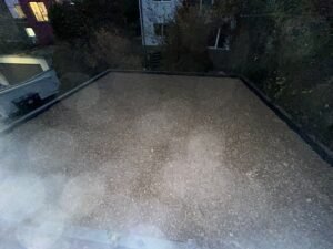 Eine rechteckig eingefasste, großflächige Kiesfläche in einem Garten bei Dämmerung oder Nacht. Die Fläche ist mit dunklen Randsteinen sauber abgegrenzt. Im Hintergrund sind dunkle Bäume, Sträucher und die beleuchteten Fenster eines benachbarten Hauses zu erkennen.