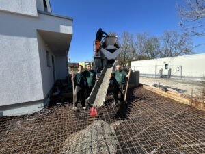 Drei Bauarbeiter in grüner Arbeitskleidung stehen lächelnd vor einem Betonmischer-LKW auf einer Baustelle. Der LKW leitet über eine Schurre frischen Beton auf eine vorbereitete Fläche mit Stahlbewehrung (Armierungsgitter). Die Arbeiten finden bei sonnigem Wetter neben einem modernen weißen Wohnhaus statt.