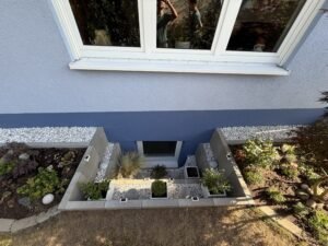 Blick von oben auf einen modern gestalteten Lichtschacht vor einem Kellerfenster. Die Einfassung besteht aus grauen L-Steinen und ist mit weißem Zierkies, kleinen Pflanzkübeln, dekorativen Steinkugeln und Solarleuchten ansprechend dekoriert. Das angrenzende Haus hat eine zweifarbige Fassade in Blau und Grau, flankiert von bepflanzten Beeten und einer weißen Kiesleiste.