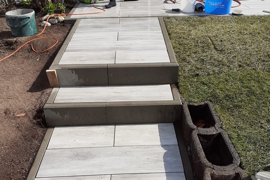 Nahaufnahme einer im Bau befindlichen Gartentreppe: Die hellen Fliesenstufen sind gesetzt, dunkle Randsteine bilden den Abschluss. Im Hintergrund Baustellen-Equipment.