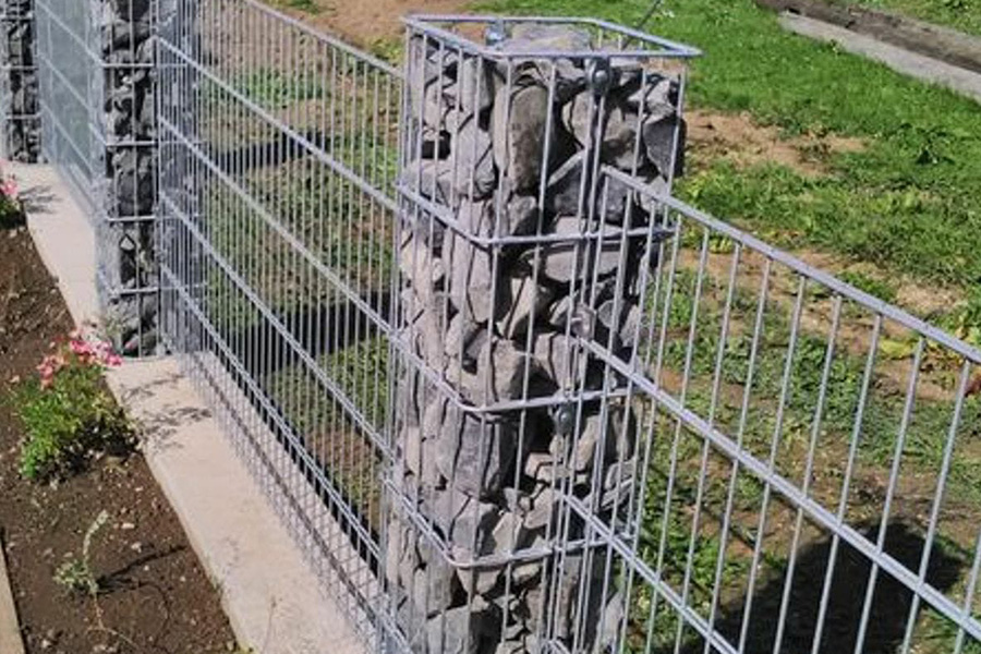 Nahaufnahme eines Zaunsystems mit einer zentralen Gabionensäule, die mit grauen Bruchsteinen gefüllt ist. Links und rechts schließen sich silberne Doppelstabmattenzäune an, die auf einem Betonfundament stehen.